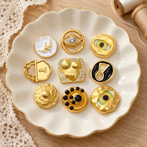 Puede incluir: Una colección de nueve botones decorativos de metal con diversas formas y diseños, incluyendo motivos de ojos con pedrería, una forma de corazón, un candado y acabados dorados texturizados, dispuestos sobre un plato de cerámica blanco.