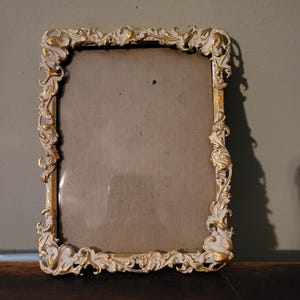May include: A rectangular picture frame featuring an ornate, raised border with a white and gold finish. The frame has intricate, swirling scrollwork details along all four sides, surrounding a plain, neutral-toned rectangular center panel.
