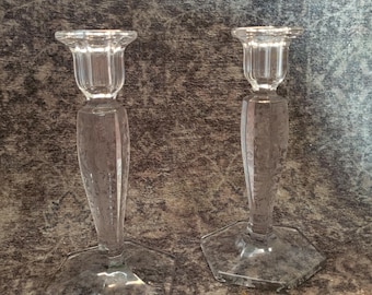Puede incluir: Un par de candelabros de vidrio transparente con una base hexagonal facetada y un tallo cónico que termina en una parte superior estriada. El vidrio es transparente y liso, diseñado para sostener una vela cónica en cada candelabro.