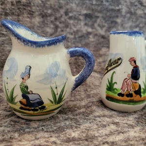 May include: Two small ceramic pitchers with a cream-colored base and blue trim. The left pitcher features a painted figure in a green top and blue skirt. The right pitcher displays a figure in a striped outfit and the text Honfleur in black script.