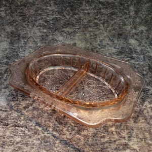 Könnte beinhalten: Eine rechteckige, unterteilte Schale aus rosa Pressglas mit abgerundeten Ecken und gewelltem Rand. Die Mitte zeigt ein geprägtes Blumenmuster, während der äußere Rand ein dekoratives Reliefdesign aufweist. Die Schale ist zweigeteilt.
