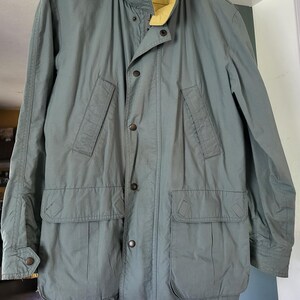 May include: A sage green jacket featuring a high collar with a bright yellow interior lining. The jacket includes a front button closure with metallic snaps, two slanted chest pockets, two large lower flap pockets, and a drawstring hem at the bottom.