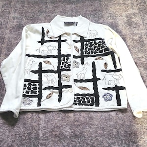 May include: A cream-coloured long-sleeve button-down shirt featuring a black grid pattern. The design includes embroidered outlines of elephants, giraffes, and camels, interspersed with black giraffe-print blocks and small decorative floral shapes.
