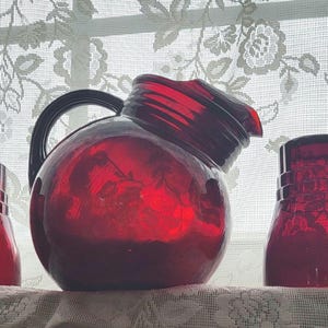 May include: A set of ruby red glassware consisting of a rounded pitcher with a handle and four matching drinking glasses. Each piece features a deep red translucent finish and a textured geometric pattern along the base of the vessels.