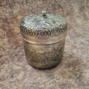 May include: A cylindrical silver-toned metal container with a removable lid. The exterior features intricate embossed floral and vine patterns, a cross-hatched textured band, and a small spherical knob centered on the top of the lid.