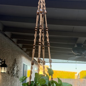 May include: A hanging plant holder made of tan macrame cord featuring a spiral knot pattern and dark brown wooden beads. The structure consists of four braided cords that converge at the top, supporting a green leafy plant in a container.