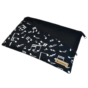 Könnte beinhalten: Schwarz-weißes Etui mit Reißverschluss zum Thema Musik. Die Tasche hat einen schwarzen Hintergrund mit einem Muster aus weißen Noten und Symbolen. Ein einfarbiges schwarzes Feld und ein braunes Etikett mit Text sind ebenfalls sichtbar.