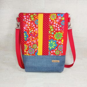 Könnte beinhalten: Eine rechteckige Umhängetasche mit einem leuchtend roten Blumenmuster aus grünen, blauen und gelben Blüten, einem zentralen gelben vertikalen Streifen und einer blauen Basis in Denim-Optik. Sie hat einen roten verstellbaren Riemen und ein braunes Etikett.