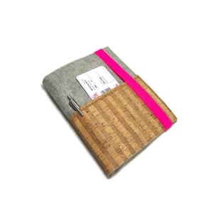 Könnte beinhalten: Ein grauer Filz-Notizbuchumschlag mit einer Kork-Vorderseite und einem leuchtend pinkfarbenen Gummibandverschluss. Ein Stift ist in einer Tasche auf der Vorderseite verstaut.