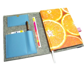 May include: A gray felt notebook cover with a blue leather interior. The cover has two pockets for cards and a pen holder. The notebook has a bright orange cover with a pattern of orange slices. The text "artista lista" is printed on a pink label.