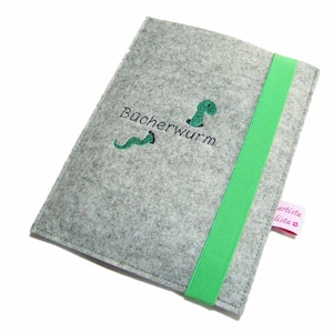 Könnte beinhalten: Graue Filz-Buchhülle mit grünem Gummizug. Die Hülle ist mit einem grünen Bücherwurm bestickt, der den Text "Bücherwurm" trägt, und einem rosa Etikett mit dem Text "artista lista"