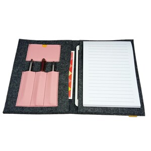 Könnte beinhalten: Offenes graues Filz-Portfolio mit einem rosa Stifthalter und einem Notizblock. Das Portfolio hat eine rosa Tasche und enthält drei Stifte, einen Stift und einen Notizblock. Der Notizblock hat liniertes Papier. Eine kleine gelbe Lasche ist sichtbar.