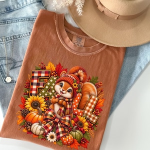 May include: A rust-colored t-shirt featuring a graphic of a squirrel wearing a knit hat and scarf, surrounded by sunflowers, pumpkins, pinecones, and autumn leaves. The word FALL is printed in large, patterned block letters behind the squirrel.