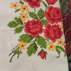 May include: A white fabric tablecloth featuring a cross-stitch pattern of large red roses, green leaves, and small yellow flowers. The floral design is arranged in a cluster on the corner of the textile, showcasing a pixelated, grid-like embroidery style.