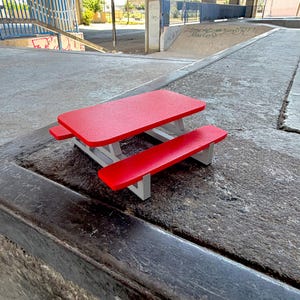 Puede incluir: Una mesa de picnic en miniatura con un tablero rectangular de color rojo brillante y dos bancos rojos a juego unidos a una estructura de color gris claro. La estructura presenta una silueta clásica de mesa de picnic con un acabado liso y una base robusta que sostiene los asientos y el tablero en un formato compacto y reducido.