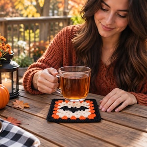 Puede incluir: Un posavasos cuadrado de ganchillo con un motivo central de murciélago negro rodeado por capas de hilo blanco y naranja, acabado con un borde negro. El posavasos está diseñado con un patrón clásico de granny square y es totalmente plano.