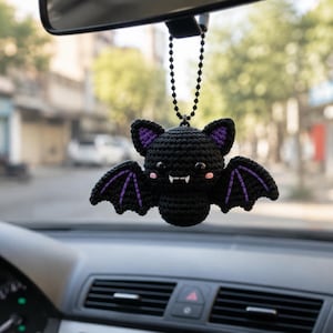 May include: A black crocheted bat hanging ornament with purple inner ear and wing details, black plastic eyes, pink stitched cheeks, and two small white fangs. The bat is suspended from a black ball chain attached to a rearview mirror.