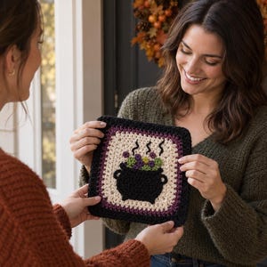 May include: A square crochet granny square featuring a black cauldron graphic in the center. The cauldron is filled with purple and green bubbling liquid with steam rising above it. The square has a cream center border and a black outer edge.