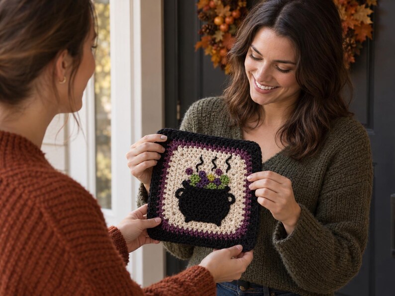 May include: A square crochet granny square featuring a black cauldron graphic in the center. The cauldron is filled with purple and green bubbling liquid with steam rising above it. The square has a cream center border and a black outer edge.