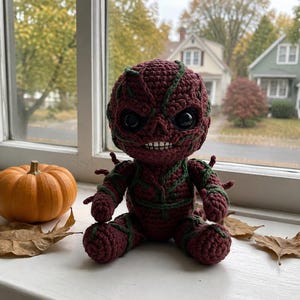 May include: A dark red crocheted doll with a textured surface and green vine-like accents. The doll features large black glossy eyes, a wide mouth with visible white teeth, and a rounded head. It is seated in a forward-facing position.