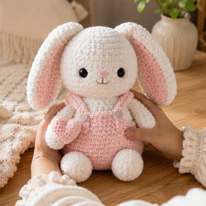 Puede incluir: Un conejo de peluche tejido a ganchillo en color blanco con orejas largas que tienen secciones interiores rosas. El conejo tiene ojos de seguridad negros, una nariz bordada en rosa y viste un peto de ganchillo rosa con dos botones plateados.