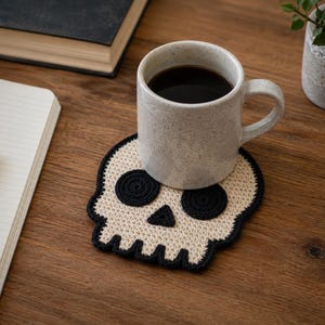 May include: A skull-shaped crochet coaster made with cream-colored yarn for the main structure and black yarn for the eye sockets, nose, and outer border. The coaster is flat and features a detailed stitched design with a defined jawline and teeth.
