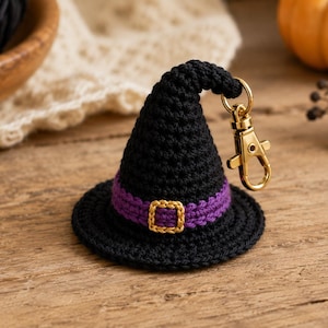 May include: A black crocheted witch hat keychain featuring a purple band around the base and a small gold-colored buckle. The hat has a pointed, slightly curved top and is attached to a gold-toned metal lobster clasp for easy attachment.