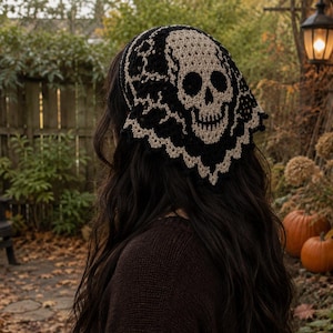 May include: A triangular crochet headscarf featuring a prominent beige skull motif centred on a black background. The accessory is crafted with intricate yarn work, showcasing a detailed skull design and a decorative scalloped lace-like border.