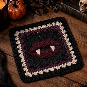 May include: A square crochet coaster featuring a dark maroon pair of lips with two white fangs centered on a black background. The design is framed by a thin purple border and an outer edge of cream-colored stitches in a decorative pattern.