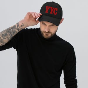 Puede incluir: Una gorra de béisbol negra con las letras YYC bordadas en rojo en la parte delantera. La gorra tiene un aspecto desgastado con una visera curva y una corona estructurada. El diseño es sencillo y llamativo, destacando el texto rojo sobre la tela negra.