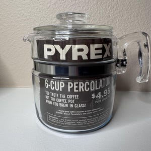 May include: A clear glass Pyrex brand 6-cup percolator with a glass lid and handle. The interior features a black paper insert with white text reading PYREX, 6-CUP PERCOLATOR, YOU TASTE THE COFFEE NOT THE COFFEE POT WHEN YOU BREW IN GLASS.