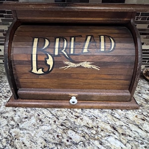 May include: A dark brown wooden bread box with a roll-top door featuring the word BREAD in large black lettering and a small gold wheat graphic. The front of the door includes a small white knob for opening and closing the storage container.