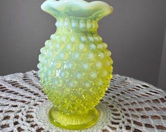 May include: A bright yellow opalescent glass vase featuring a hobnail texture across its bulbous body. The vase has a flared, ruffled rim and sits on a circular pedestal base, showcasing a translucent quality that shifts in color intensity.