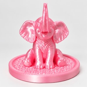 May include: A shiny, bright pink elephant figurine in a seated position with its trunk raised upward. The elephant is attached to a circular, textured pink base. The entire object features a smooth, reflective finish with visible 3D print lines.