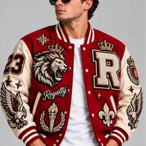 Pode incluir: Uma jaqueta varsity vermelha e creme com patches bordados de um leão com coroa, a letra R, águias, uma tocha e a palavra Royalty. A jaqueta tem fecho de botões de pressão, punhos canelados listrados e uma camiseta branca por baixo.