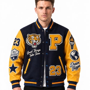 Op de afbeelding: Een marineblauw en geel varsity-jack met een tijgerkop-patch, de letter P, het getal 23 en de tekst Good Things Take Time, Built Different en Dream Bigger. De mouwen zijn geel met diverse patches en hebben blauwe boorden.