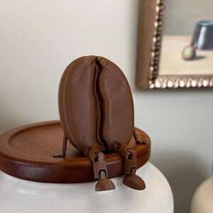 May include: A brown coffee bean figurine with articulated limbs sitting on the wooden lid of a white ceramic canister. The canister features a rectangular label with the text 2LB/32 OZ embossed on the front, indicating its capacity.