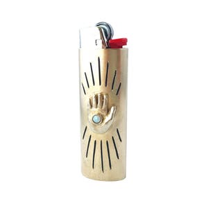 May include: Gold lighter with a hand design and a blue stone in the palm. The lighter has a black and white sunburst design around the hand.