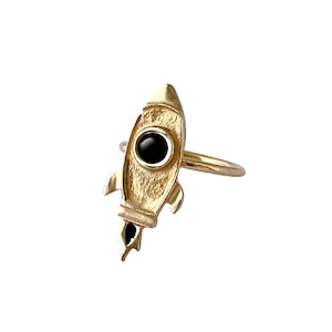 Könnte beinhalten: Ein goldfarbener Raketenring mit einem schwarzen Stein in der Mitte. Der Ring ist so gestaltet, dass er wie eine Rakete mit einer spitzen Nase und einer Flamme aussieht, die aus dem Heck kommt.