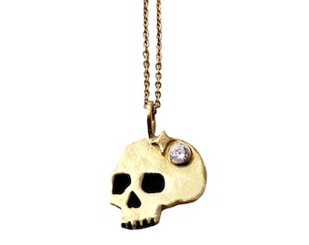 Könnte beinhalten: Eine goldene Totenkopf-Halskette mit einem kleinen Diamanten, der in die Stirn des Schädels eingefasst ist. Die Kette ist eine einfache goldene Kette.