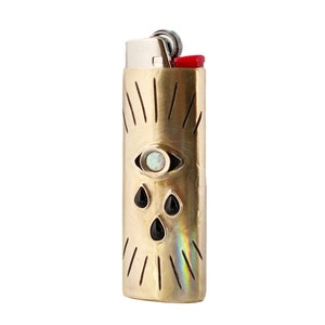 May include: A gold-colored lighter case with an eye and teardrop design. The eye features an opal-like center, and the teardrops are black. The top of the lighter has a red button.