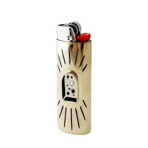May include: A gold lighter with a black and white design of a window with stars and a moon. The lighter has a red top and a silver hinge.