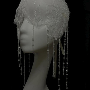 May include: A white lace headpiece featuring a delicate floral pattern and scalloped edges. The cap is adorned with numerous dangling strands of clear, faceted beads that hang down from the lace base, creating a fringe effect around the head.