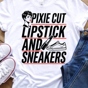 Peut inclure: Un t-shirt blanc avec une impression graphique noire portant le texte PIXIE CUT, LIPSTICK, AND SNEAKERS. Le motif comprend une illustration stylisée d'une personne aux cheveux courts, un tube de rouge à lèvres et une basket, séparés par des lignes rouges.