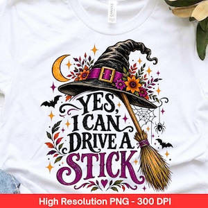 Può includere: Una t-shirt bianca con una grafica raffigurante un cappello da strega nero con fascia viola, una scopa, una luna crescente gialla, ragnatele, pipistrelli e fiori colorati. Il testo recita "YES, I CAN DRIVE A STICK" in caratteri neri e viola.
