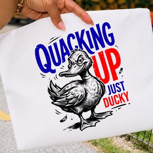 Peut inclure: Une pochette en tissu blanc avec une illustration en noir et blanc d'un canard à l'expression malicieuse. Le texte indique QUACKING UP JUST DUCKY en lettres bleues et rouges. Le motif comprend de petites plumes graphiques éparpillées.
