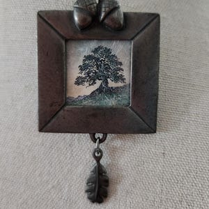 May include: A square metal brooch featuring a dark, oxidized finish with two sculpted acorns at the top. The center contains a small, detailed illustration of a tree on a hill. A single metal leaf charm hangs from the bottom of the frame.