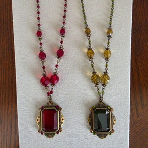 May include: Two necklaces featuring rectangular pendants in red and dark green, each set in an ornate bronze-toned metal frame. The chains are composed of matching red or green translucent beads interspersed with small bronze-toned metal spacers.