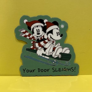 May include: A die-cut sticker featuring Mickey and Minnie Mouse wearing red and white Santa hats and matching scarves while riding a dark green sled. The background is a muted green with a small white snowflake and the text Your Door SLEIGHS!