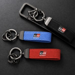 May include: Three keychains featuring a metal ring and a suede-like strap in black, blue, and red. Each strap displays the GR SPORT logo in white and red text. The black keychain includes a rectangular metal frame attachment with additional rings.